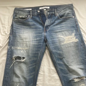 Blå slitna jeans från Calvin Klein. Pris går att sänka  - Säljer ett par blå jeans från Calvin Klein med snygga slitningar och hål på båda benen. Jeansen har klassisk femficksdesign, raka ben och tydliga slitdetaljer för en cool, avslappnad look. Perfekta för dig som gillar streetstyle.