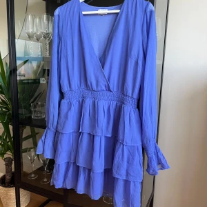 Blå omlottklänning från Bubbleroom - Supersöt blå omlottklänning från Bubbleroom med volanger och smock i midjan. Klänningen har långärmade ärmar med smock vid ärmslut och v-ringad hals. Perfekt för dig som gillar en flowy och feminin stil.