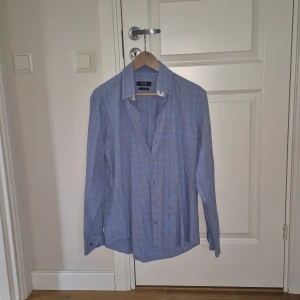 Blårutig skjorta från Bugatti, M - Snygg blårutig skjorta från Bugatti i storlek M. Skjortan har ett klassiskt krage, långa ärmar och knappar framtill. Materialet är mjuk bomull och mönstret är rutigt med tunna röda linjer. Insidan av knappslån har ett diskret blommigt tyg för extra stil.