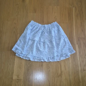 Blommig volangkjol från Primark - Supersöt kort kjol från Primark med ljusblått blommönster på vit botten. Kjolen har volanger och resår i midjan med snörning för extra comfy passform. Perfekt för sommarens alla dagar, lätt och luftig i bomull.