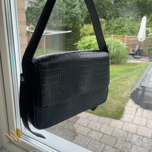 Svart axelväska från Bik Bok - Snygg svart axelväska från Bik Bok med croco-mönstrad yta och bred axelrem. Väskan har magnetlås och ett stort fack med innerficka med dragkedja. Perfekt för att bära dina viktigaste grejer med stil.