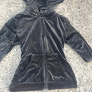 Mörkgrå velour hoodiejacka - Mysig hoodiejacka i mörkgrå velour med dragkedja framtill och stora fickor. Jackan har huva med metallöljetter och långa ärmar. Perfekt för en chill och avslappnad stil.