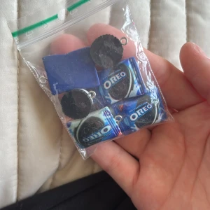 Oreo-inspirerade smyckesberlocker - Säljer ett gäng handgjorda smyckesberlocker med Oreo-tema! Paketet innehåller små berlocker formade som Oreo-kakor, blå och vita rektanglar samt miniatyrer av Oreo-förpackningar. Perfekt att använda till egna halsband, armband eller örhängen. Färgerna är svart, blå och vit.