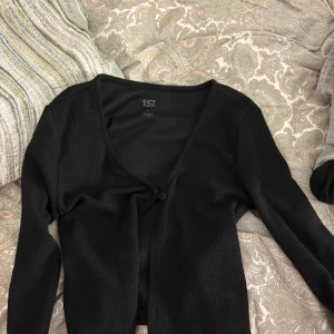 Svart ribbad kofta från 157 - Svart ribbad kofta från 157 med v-ringning och en knapp framtill. Långärmad och i mjukt, stretchigt material som sitter snyggt på kroppen. Perfekt att slänga över en topp eller klänning för en chill look.