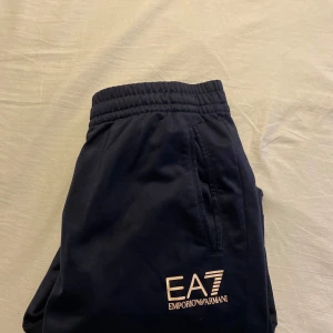 EA7 Emporio Armani svarta mjukisbyxor - Snygga mörkblåa mjukisbyxor från EA7 Emporio Armani med elastisk midja och muddar vid bensluten. Byxorna har fickor på sidorna och tydlig logga framtill. Perfekta för chill eller träning, med sportig och clean look.nypris 888kr