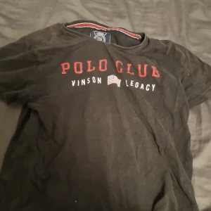 Svart Polo Club t-shirt med tryck - Svart t-shirt från Vinson Polo Club med rött och vitt tryck på bröstet där det står 'POLO CLUB VINSON LEGACY' och en liten amerikansk flagga. Rund hals med röd och vit detalj på insidan. Klassisk passform och korta ärmar, perfekt för en chill look.