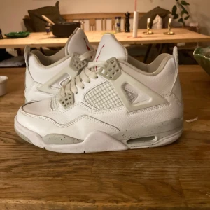 Nike Air Jordan 4 Retro vit - Säljer ett par Nike Air Jordan 4 Retro i helvitt skinn med meshpaneler och grå detaljer på sulan. Skorna har klassisk snörning, Jumpman-logga i rött på plösen och bak på hälen. Perfekt för dig som vill ha en clean och ikonisk sneaker med snygga detaljer.