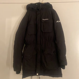 Svart jacka från Everest, stl 34 - Svart jacka från Everest med huva och flera stora fickor framtill. Jackan har dragsko i midjan för snygg passform och stängs med dragkedja och knappar. Perfekt för kalla dagar med sin långa modell och varma vaddering.
