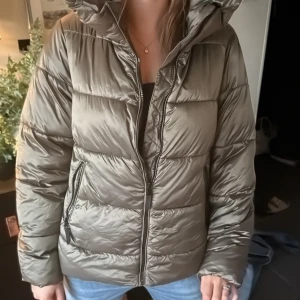 Olivgrön pufferjacka från Vero Moda - Säljer en olivgrön pufferjacka från Vero Moda med huva och dragkedja framtill. Jackan har quiltad design, två fickor med dragkedja och är perfekt för kyliga dagar. Materialet är syntet och jackan har en normal passform med långa ärmar.