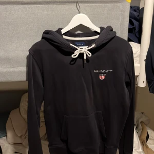 Mörkblå hoodie från GANT med logga - Svart hoodie från GANT i storlek M med broderad logga och emblem på bröstet. Tröjan har vit snörning i huvan, känguruficka och mjukt material som känns skönt mot huden. Perfekt för chill dagar och enkel att matcha med jeans eller joggers.