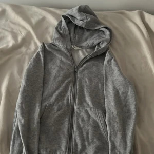 Grå hoodie med dragkedja från H&M - Enkel och stilren grå hoodie från H&M i storlek 146/152. Tröjan har huva, hel dragkedja framtill och två fickor. Perfekt för chill dagar eller när du vill ha något bekvämt och basic. Materialet är mjukt och skönt, troligtvis bomullsmix.