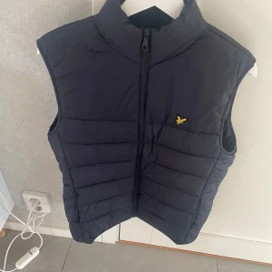 Mörkblå dunväst från Lyle & Scott - Snygg mörkblå dunväst från Lyle & Scott med klassisk gul logga på bröstet. Västen har hög krage, quiltad design och dragkedja framtill. Perfekt att slänga över en hoodie eller tröja för extra värme.