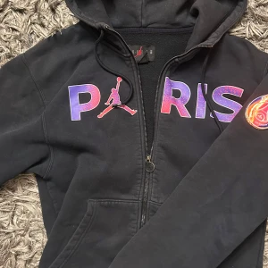 Jordan PSG svart hoodiejacka XS - Svart hoodiejacka från Jordan x Paris Saint-Germain med dragkedja, huva och färgstarkt PARIS-tryck i lila och rosa på bröstet. PSG-märke på ärmen och mjukt fleecefoder. Perfekt för dig som gillar sportig streetstyle.