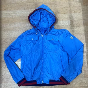 Blå vindjacka windbreaker från Moncler med huva - Snygg blå vindjacka från Moncler med huva och dragkedja framtill. Jackan har två bröstfickor med knappar, ribbade muddar i blått och vinrött samt Moncler-logga på ärmen. Perfekt för dig som vill ha en stilren och sportig look.