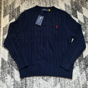 Mörkblå kabelstickad tröja Polo Ralph Lauren - Kabelstickad mörkblå tröja från Polo Ralph Lauren med rund halsringning och röd broderad logga på bröstet. Tröjan har långa ärmar och ribbade muddar för en klassisk look. Perfekt för dig som gillar stilren och tidlös design.