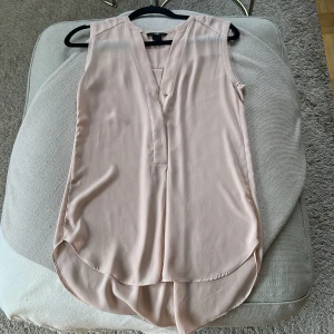 Ljusrosa ärmlös blus från H&M - Säljer en ljusrosa ärmlös blus från H&M i flowy och mjukt material. Blusen har v-ringning framtill, längre rundad nederkant och en stilren look som passar perfekt till jeans eller kjol. Enkel och snygg design som är lätt att matcha.