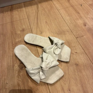 Vita sandaler med rosett - Snygga vita sandaler med fyrkantig tå och stora rosetter framtill. Perfekta för sommaren och ger en trendig touch till din outfit. Gjorda i syntetmaterial och har platt sula för extra komfort.