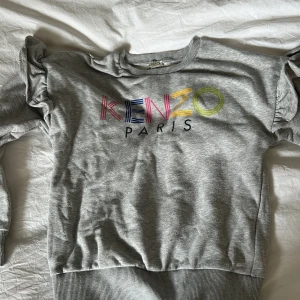 Grå Kenzo sweatshirt med volang - Säljer en grå sweatshirt från Kenzo med färgglatt logotryck på bröstet. Tröjan har långa ärmar med snygga volangdetaljer vid axlarna och ribbade muddar vid ärmslut och nederkant. Perfekt för dig som gillar streetstyle och vill sticka ut med coola detaljer.