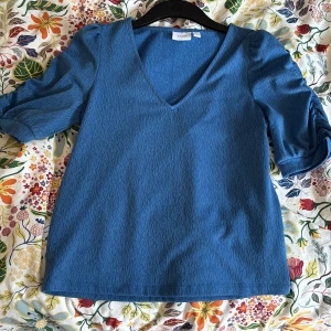Blå blus med puffärm från Vila - Snygg blå blus från Vila med v-ringning och puffiga korta ärmar med rynkdetalj. Materialet har en lätt struktur som ger en cool känsla. Perfekt för dig som gillar stilrena plagg med lite edge. Passar dig som vill ha en blus som sticker ut utan att vara för mycket.