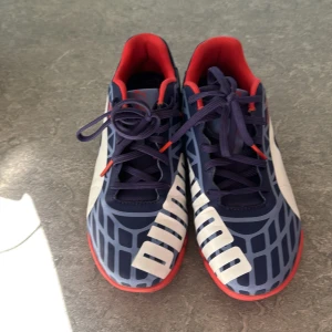 Blå och röda Puma skor  - Säljer ett par snygga Puma skor i mörkblå med röda detaljer och vit logga. Skorna har ett grafiskt mönster, snörning och platt sula. Insidan är röd och yttermaterialet är syntet. Själva överdelen och insidan av skon i mycket fint skick .Lätta i vikt. Barnen vill sälja skorna därav priset.