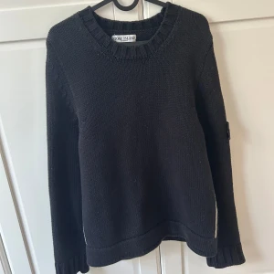 Svart stickad tröja Stone Island - Unik Svart stickad tröja från Stone Island med klassisk rund hals och ribbade muddar. Tröjan har den ikoniska Stone Island-loggan på vänster ärm och är tillverkad i mjuk bomull med elastan för extra komfort. Perfekt för dig som gillar streetwear och snygga detaljer.