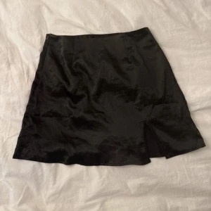 Svart kortkjol med slits - Snygg svart kortkjol med lätt glansig yta och en liten slits framtill för extra edge. Kjolen har en enkel, rak form och är perfekt för dig som gillar minimalistisk stil. Materialet känns mjukt och har en silkesliknande finish.