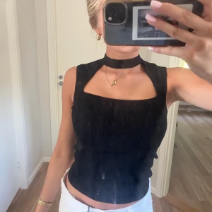 Svart ribbad ärmlös topp med choker - Säljer en svart ribbad topp med fyrkantig urringning och inbyggd choker-detalj runt halsen. Toppen är ärmlös och har en tight passform som framhäver siluetten. Perfekt för dig som gillar trendiga och stilrena plagg.