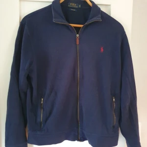 Mörkblå ziptröja Polo Ralph Lauren - Mörkblå ziptröja från Polo Ralph Lauren med klassisk krage och två fickor med dragkedja. Jackan har röd broderad logga på bröstet och är tillverkad i bomull. Perfekt för dig som gillar stilren och enkel design.