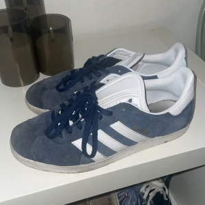 Adidas Gazelle blå sneakers mocka - Snygga Adidas Gazelle sneakers i mörkblå mocka med vita detaljer och klassiska tre ränder på sidan. Skorna har snörning och låg profil, perfekt för en clean och sportig look. Guldtexten 'Gazelle' på sidan ger extra stilpoäng. Skorna är väll andvända, dom är i acceptabelt skick skulle jag säga 👍🏼