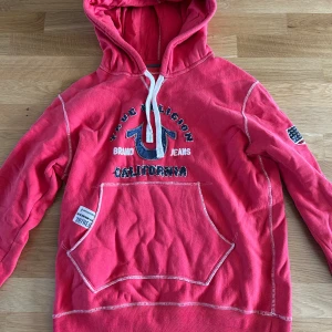 Röd hoodie från True Religion - Säljer en röd hoodie från True Religion med broderad logga och text på bröstet, samt USA-flagga på ärmen. Tröjan har vit snörning i huvan, stor magficka och kontrastsömmar. Materialet är mjuk bomull, perfekt för chill dagar. Passformen är normal och ärmarna är långa.