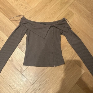 Brun ribbad offshoulder topp - Säljer en brun ribbad offshoulder topp från Gina Tricot med långa ärmar och dekorativa knappar framtill. Toppen har en snygg asymmetrisk krage och smala axelband som ger en unik look. Perfekt för dig som gillar trendiga detaljer och stilren design.