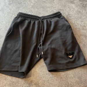 Svarta shorts från Emporio Armani - Svarta shorts från Emporio Armani med elastisk midja och snörning. Mjukt bomullsmaterial och klassisk passform. På vänster ben finns en svart och guldig logotypdetalj. Perfekta för en chill och stilren look.