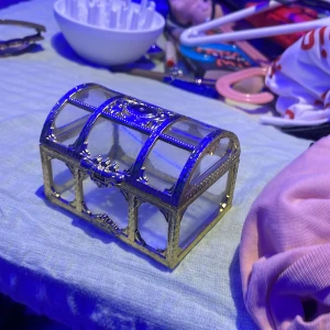 Glasad smyckesask med guldfärgade detaljer - Säljer en elegant smyckesask i glas med guldfärgade metallkanter och dekorativa mönster. Asken har en kupolformad öppningsbar lock och genomskinliga sidor som visar dina smycken snyggt. Perfekt för att förvara ringar, halsband eller örhängen på ett stilrent sätt.