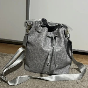 Grå axelväska från Tamaris - Snygg grå axelväska från Tamaris med justerbar axelrem och coolt mönster med logotypen över hela väskan. Väskan har dragskostängning och detaljer i silver. Perfekt för dig som vill ha en stilren och trendig väska med mycket plats.