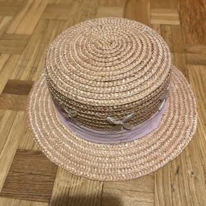 Rosa stråhatt från H&M med band - Söt rosa stråhatt från H&M med brett, ljusrosa band runt kullen och dekorativa blommor i transparent material. Klassisk form med platt kulle och brätte, perfekt för soliga dagar och sommarhäng.