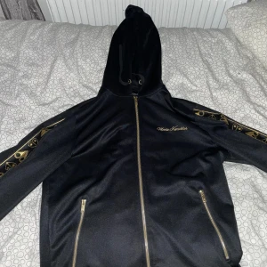 Svart Moose Knuckles kofta med huva - Svart kofta från Moose Knuckles med huva och guldfärgade detaljer. Dragkedja framtill och två fickor med guldfärgade dragkedjor. Ärmarna har ett snyggt guldmönster längs sidorna och logga broderad på bröstet. Perfekt för dig som gillar streetwear med exklusiv känsla.