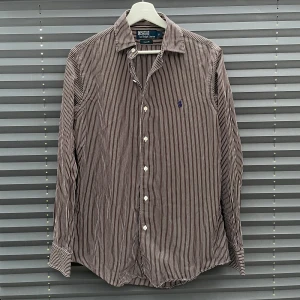 Ralph Lauren skjorta - Snygg brun skjorta från Polo Ralph Lauren i storlek S med vita smala ränder och klassisk krage. Skjortan har lång ärm, knäppning framtill och broderad blå logga på bröstet. Kom dm vid minsta fråga eller fundering och ♦️kom gärna med bud! ♦️