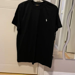 Svart t-shirt från Ralph Lauren - Klassisk svart t-shirt från Polo Ralph Lauren med vit broderad logga på bröstet. Enkel och stilren design med rund hals och korta ärmar. Perfekt basplagg för en clean look.