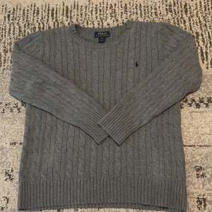 Grå stickad tröja från Polo Ralph Lauren - Säljer en snygg grå kabelstickad tröja från Polo Ralph Lauren med klassisk rund hals och långärmad passform. Tröjan har det ikoniska Polo-logot broderat på bröstet och är gjord i mjuk stickad bomull. Perfekt för dig som gillar stilrena och tidlösa plagg.