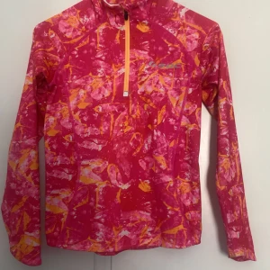 Färgstark träningshoodie från SOC - Säljer en långärmad hoodie från SOC med färgstarkt abstrakt mönster i rosa, orange och vitt. Tröjan har half zip framtill och är tillverkad i ett lätt och stretchigt syntetmaterial, perfekt för träning eller utomhusaktiviteter.