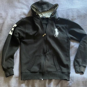 Svart hoodie från Polo Ralph Lauren - Svart hoodie med dragkedja från Polo Ralph Lauren. Stor vit logga på bröstet och en stor siffra 3 på ena ärmen. Huva med knappar och fickor framtill. 