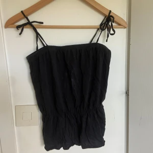 Svart linnetopp med smala axelband från Size 8 Needle - Svart linnetopp med smala knytbara axelband från Size 8 Needle. Toppen är tillverkad i en mjuk blandning av bomull och modal, har en avslappnad passform och resår i midjan för en bekväm känsla.