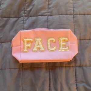Rosa necessär med glittrig text - Säljer en rosa necessär med stora bokstäver i guldglitter som stavar 'FACE' på framsidan. Väskan har dragkedja med guldfärgad detalj och är rymlig inuti. Perfekt för smink eller småprylar.