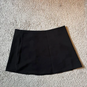 Svart kortkjol - Enkel och stilren svart kortkjol med rak passform. Kjolen är utan synliga detaljer och passar perfekt till många olika outfits. Materialet känns mjukt och följsamt. och även en dragkedja på sidan