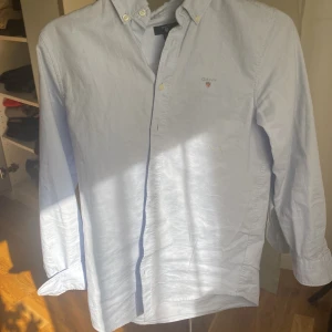 Ljusblå skjorta från GANT - Klassisk ljusblå skjorta från GANT med broderad logga på bröstet och knappar framtill. Skjortan har button-down krage och långa ärmar. Perfekt för en stilren och tidlös look. 11-12 år