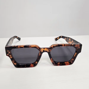 Bruna fyrkantiga solglasögon med leopardmönster - Snygga solglasögon med kraftig fyrkantig båge i brunt leopardmönster och mörka glas. Perfekt accessoar för att sticka ut och ge en cool vibe till din outfit.