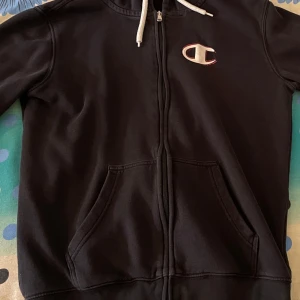 Svart hoodie med dragkedja från Champion - Säljer en svart hoodie från Champion med dragkedja och vit dragsko. Tröjan har två fickor framtill och ett broderat Champion-märke på bröstet. Materialet är 78% bomull och 22% polyester.