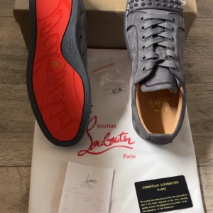 Christian Louboutin - Säljer ett par gråa sneakers från Christian Louboutin med ikoniska röda sulor och grå nitar på tån. Skorna har snörning och är tillverkade i mocka med läderfoder. Perfekta för dig som vill sticka ut med en lyxig och edgy stil.