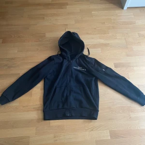 Svart hoodie från Peak Performance - Svart hoodie med dragkedja från Peak Performance. Tröjan har huva med dragsko och vit logga på bröstet. Perfekt för en avslappnad stil och enkel att matcha med andra plagg.