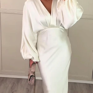 Vit långklänning med v-ringning och ballongärmar - Elegant vit långklänning med djup v-ringning och vida ballongärmar. Klänningen har en figurnära midja och är tillverkad i ett slätt, glansigt material som ger en lyxig känsla.